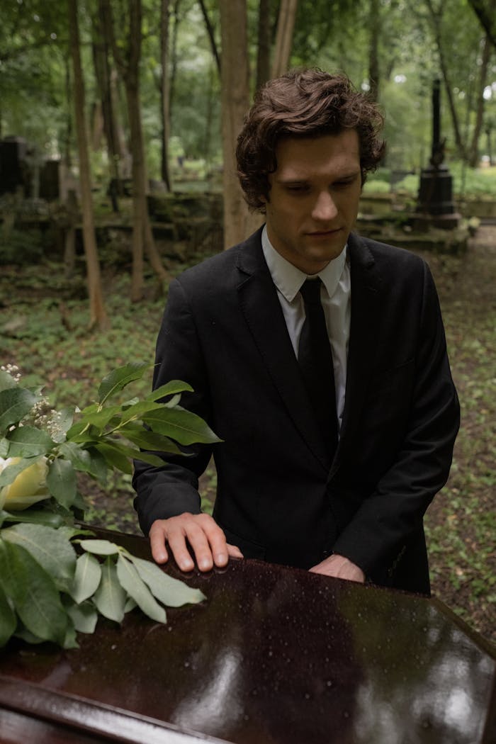 A solemn man in a suit stands by a coffin at an outdoor funeral, surrounded by greenery.