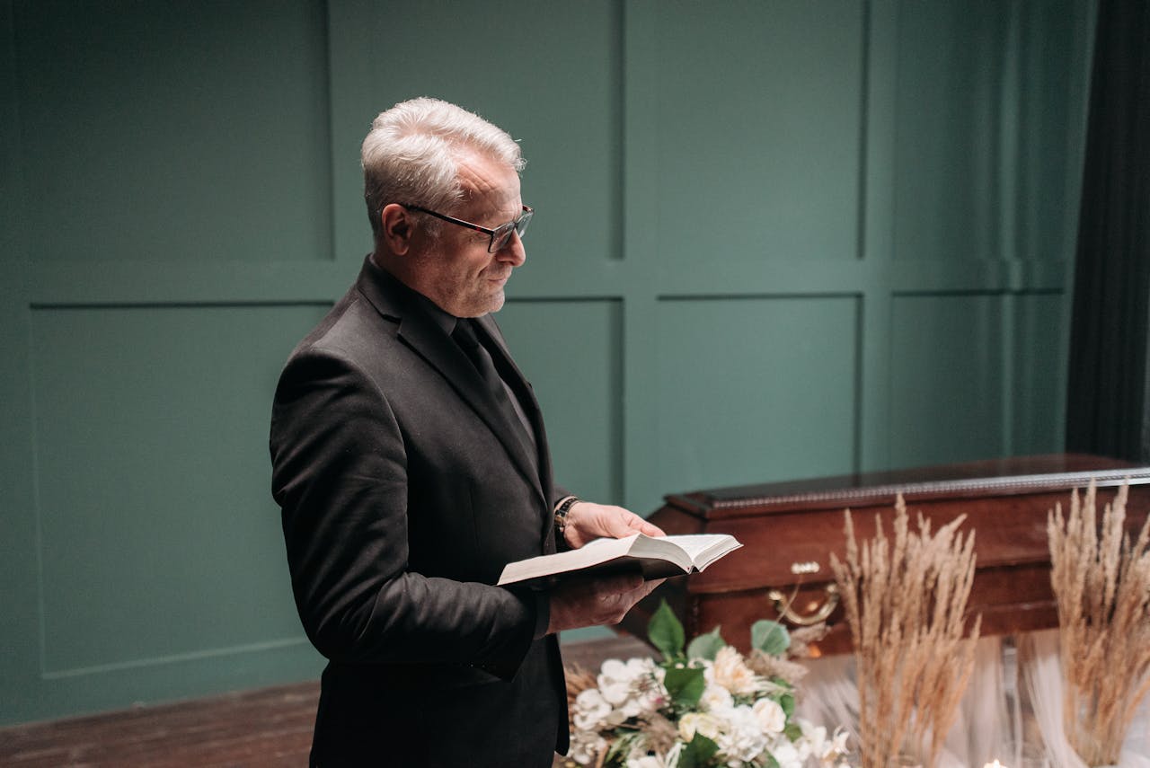 A pastor reads from the Bible during an indoor funeral service with a closed casket and floral arrangements.