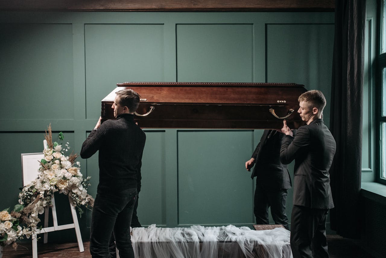 Pallbearers carry a wooden coffin at an indoors funeral service, surrounded by floral tributes.