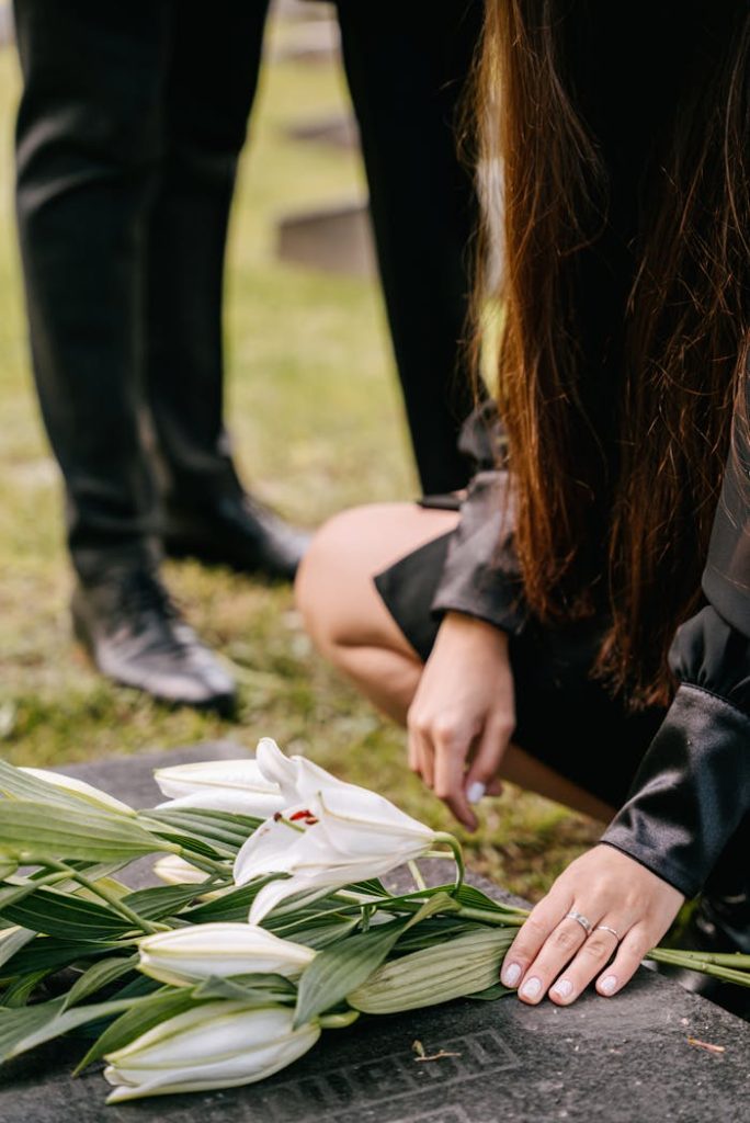 A woman kneels by a grave adorned with white lilies, symbolizing loss and remembrance.